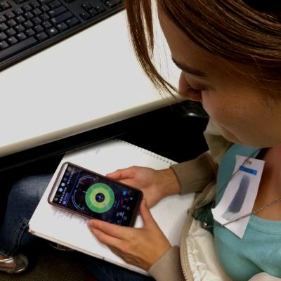 A person sitting at a desk looks at a smartphone displaying a colorful circular radar-like interface, possibly related to GPS or satellite tracking. The individual holds the phone over a notepad and wears a visitor badge with a blurred name tag. A computer keyboard is visible on the desk nearby.