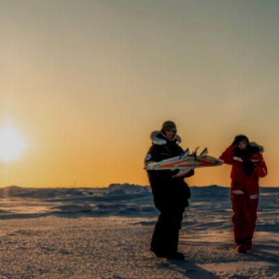 Two people wearing heavy winter gear stand on a snowy landscape at sunset. One person, dressed in black, is holding a guitar, while the other, dressed in red, is listening and smiling. The sky is clear with the sun low on the horizon, casting a warm golden glow across the icy terrain