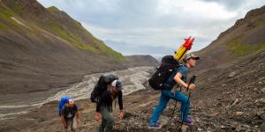 Three field crew members hiking up the mountain with glacier depicted in the mountain valley below them