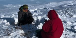 Two people sitting together on a snow pack field, one person filming the other talk