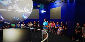 A presenter stands in front of a large illuminated spherical display showing Earth, addressing a seated audience in a dark blue-walled room. The setting resembles an interactive science exhibit or educational presentation, likely featuring real-time Earth data or visualizations.