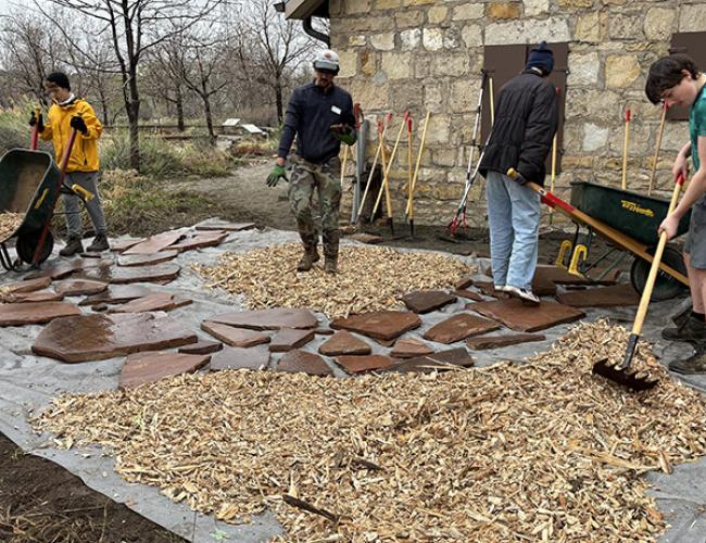 Four people working on a walkway in front of a stone house