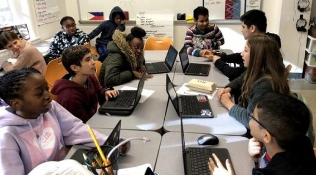 A group of students using laptops at desks all pushed together to form one collaborative workspace.