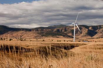 A white wind turbine turns in front of green and tan mountains. There are dried tall grasses in the foreground.