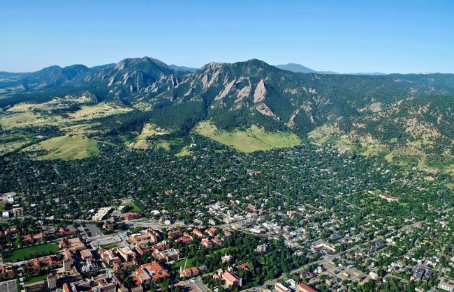 Aerial view of Boulder, Colorado, showing the city with its grid-like streets surrounded by lush greenery, with the Flatirons rock formations and mountains in the background under a clear blue sky