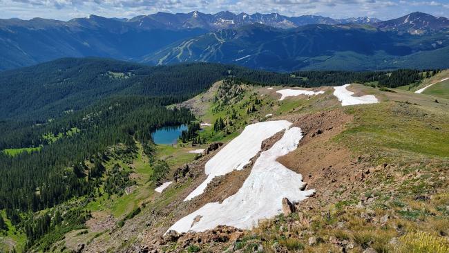 A scenic mountain landscape with patches of melting snow on a rocky slope in the foreground. Below, a small turquoise alpine lake is surrounded by dense evergreen forest. In the distance, tall rugged peaks rise above the tree line under a partly cloudy sky