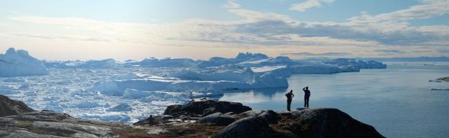Landscape shot of Greenland ice sheets in the background and rocks in the foreground. Two polar explorers stand on top of the rocks gazing out into the polar sheets.