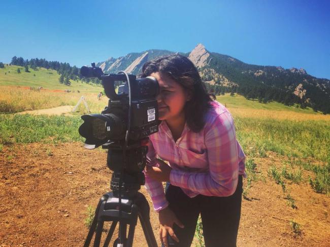 Person in a pink plaid shirt operating a video camera on a tripod in an open field with mountains in the background under a clear blue sky.
