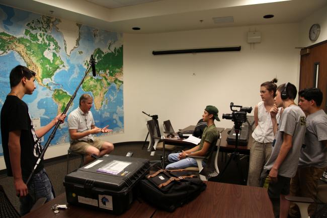 Group of students filming an interview indoors; one person holds a boom microphone, another operates a camera, while a man sits in front of them speaking, with a large world map on the wall behind